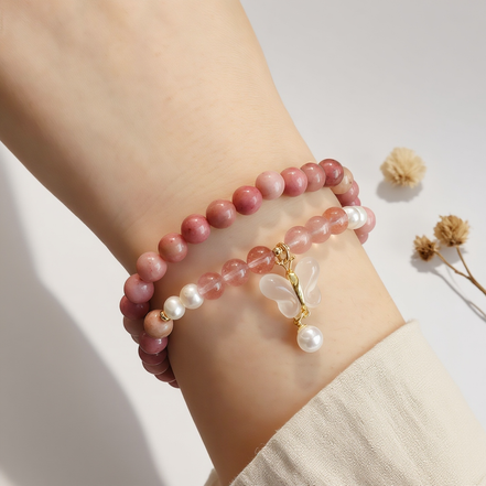 Double-layer pink gemstone bracelet with pearls and a butterfly charm, displayed on soft cream-colored fabric in natural daylight.